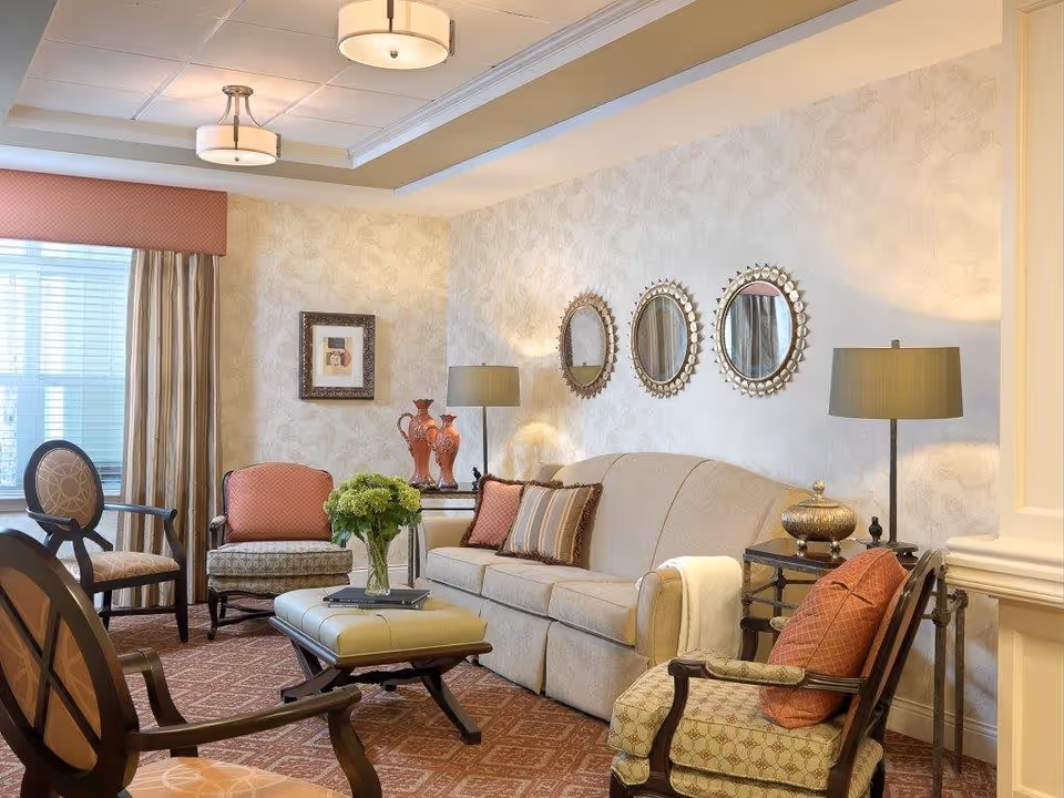 A cozy living room with a beige sofa adorned with decorative pillows, two armchairs with patterned cushions, a cushioned ottoman with a vase of green flowers on top, two side tables with lamps and decorative items, three round mirrors on the wall, and a window with curtains allowing natural light to enter.