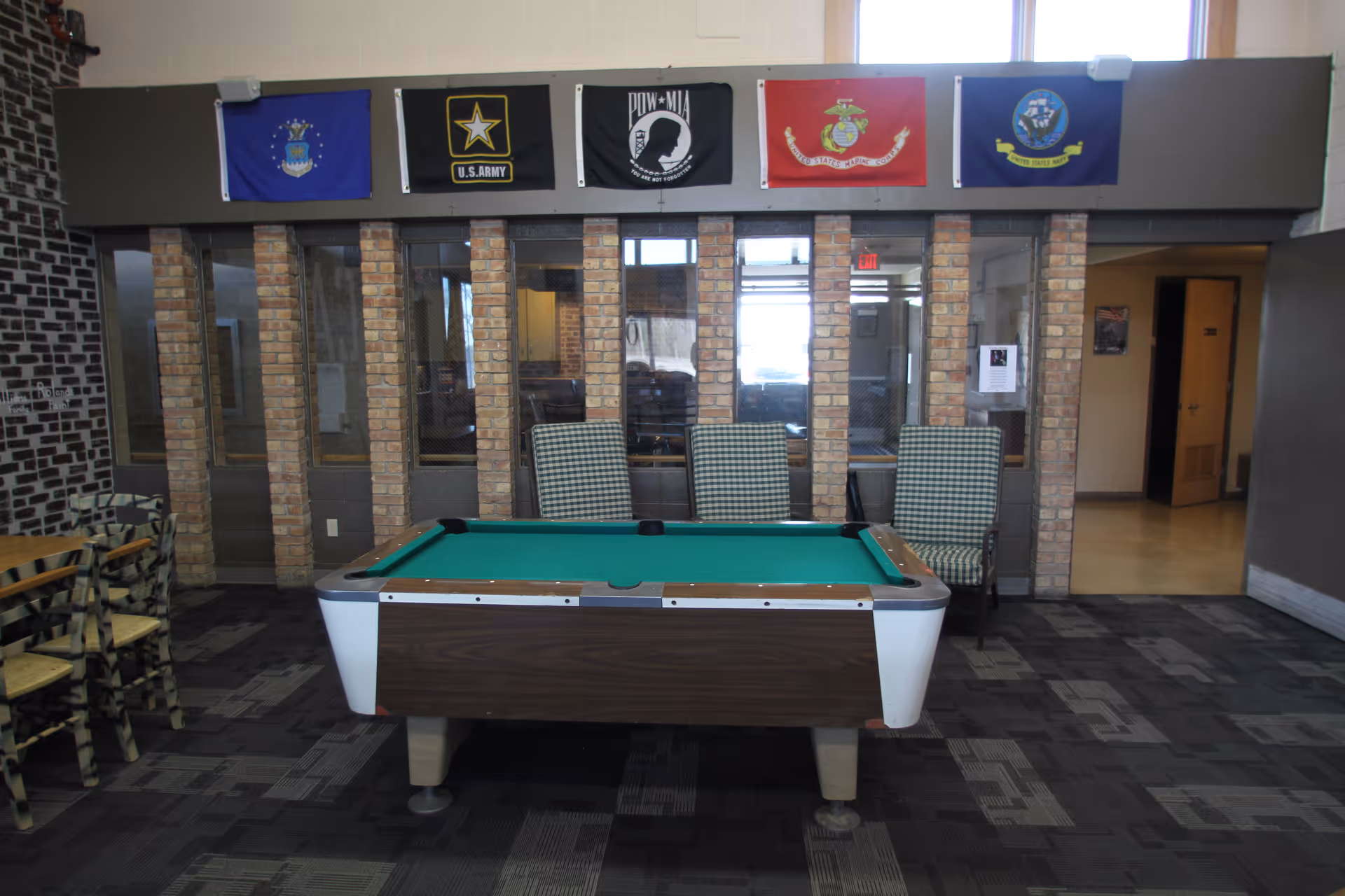 A pool table sits in a senior community lounge area with chairs in front of a glass-partitioned wall and military flags hanging above.