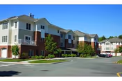 Exterior view of a multi-story residential building with balconies, surrounded by trees and a parking area under a clear blue sky.