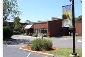 Single-story brick building with a paved parking lot, landscaped shrubs, and a lamppost banner featuring a sunflower.