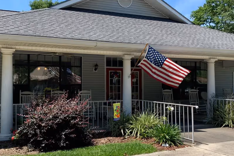 Front porch of a building with white columns, a ramp with metal railings, an American flag mounted on one of the columns, rocking chairs on the porch, and landscaping with bushes and grass in front.