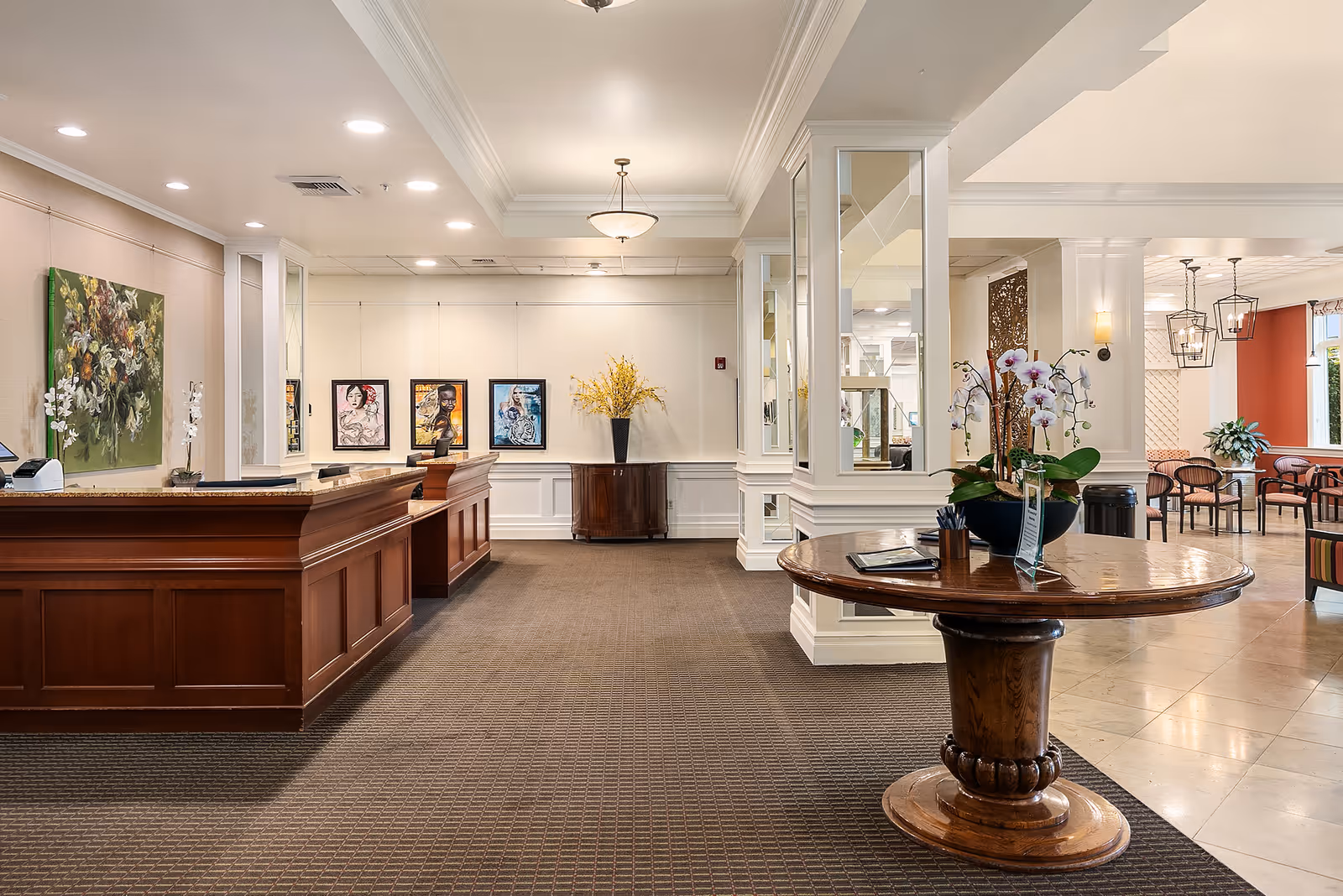 Bright senior living lobby with a wooden reception desk, round table topped with orchids, and seating area beyond.