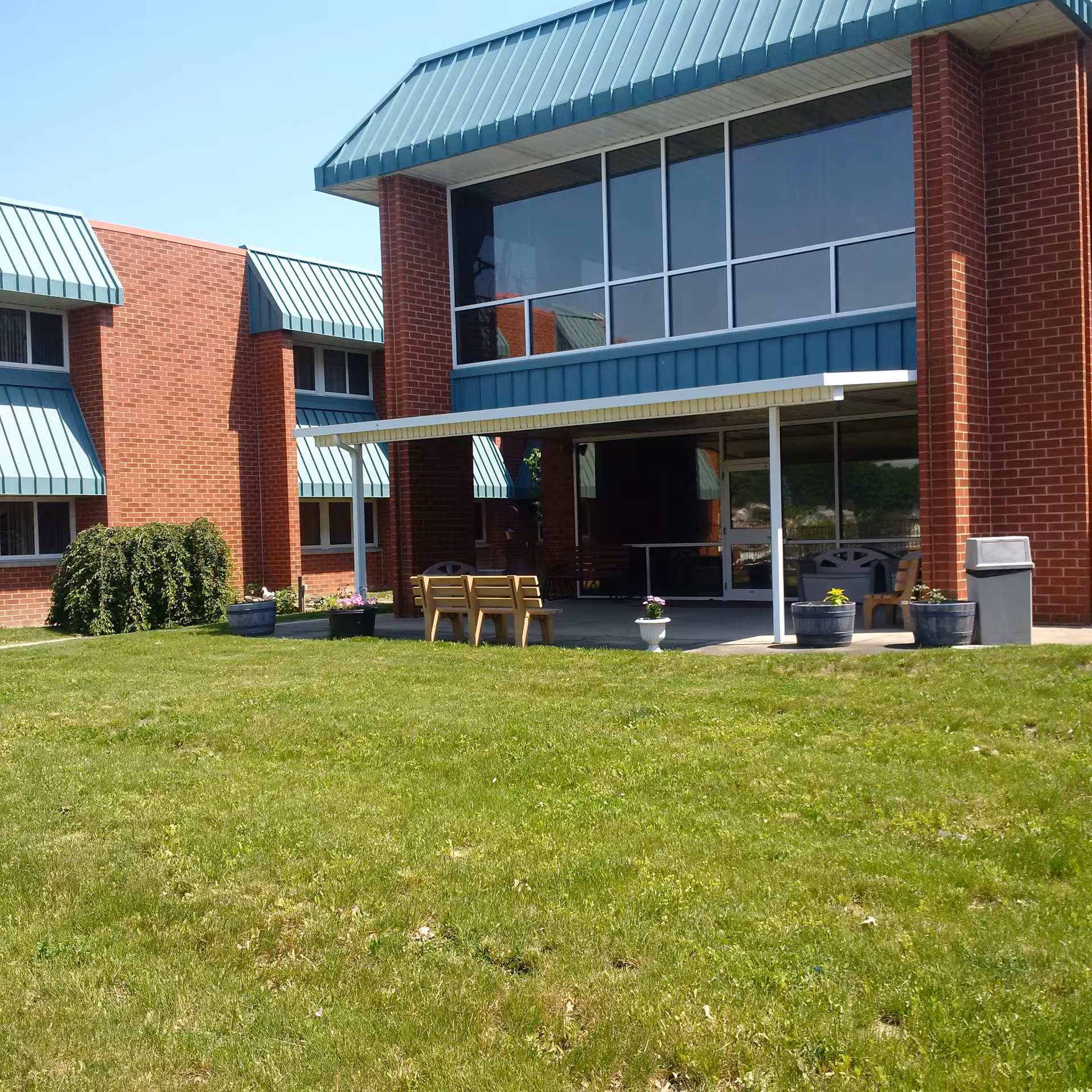 Outdoor patio area of a brick building with large windows and teal metal awnings. The patio has a covered section with wooden benches, potted plants, and a trash can. The foreground shows a well-maintained grassy lawn under a clear blue sky.