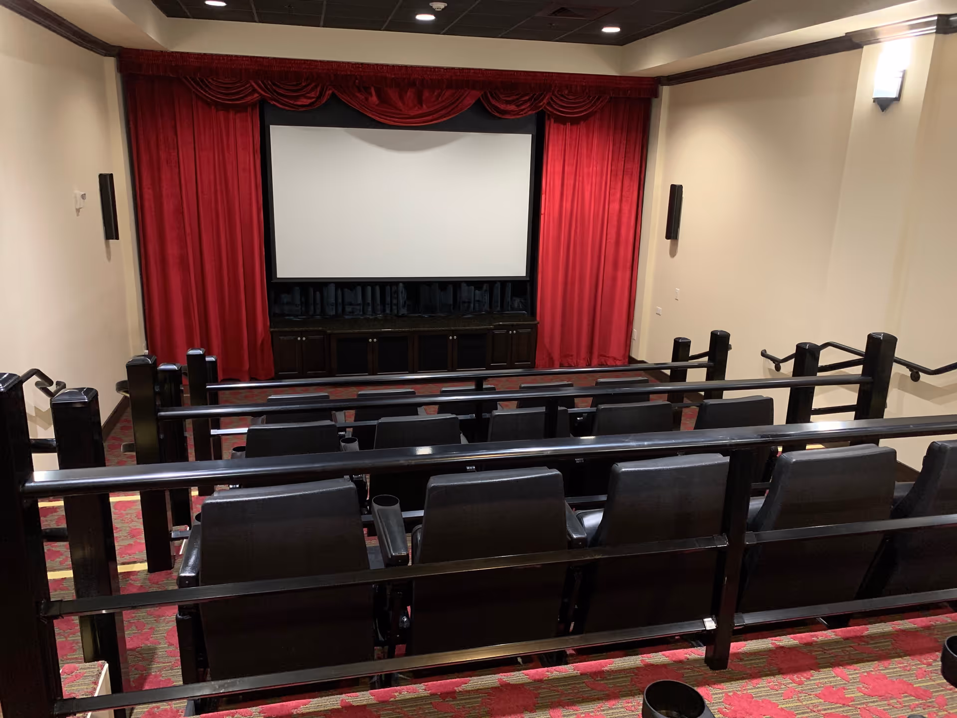 A small theater room with rows of black seats facing a large white projection screen framed by red curtains. The room has beige walls, dark wood railings, and red patterned carpet.