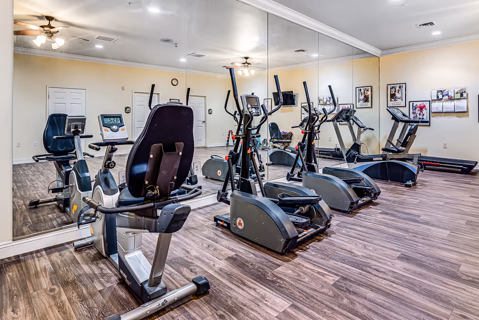 A senior living facility fitness room with exercise equipment including recumbent bikes, elliptical machines, and a treadmill. The room has wood flooring, beige walls, a large wall mirror, ceiling fans with lights, and framed pictures on the wall.