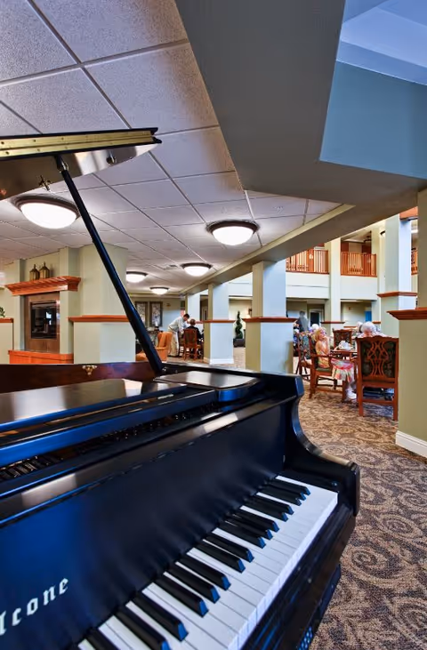 Interior view of a senior living community common area featuring a black grand piano in the foreground. In the background, several elderly residents are seated at tables engaged in conversation. The space has carpeted floors, columns, and a high ceiling with recessed lighting.