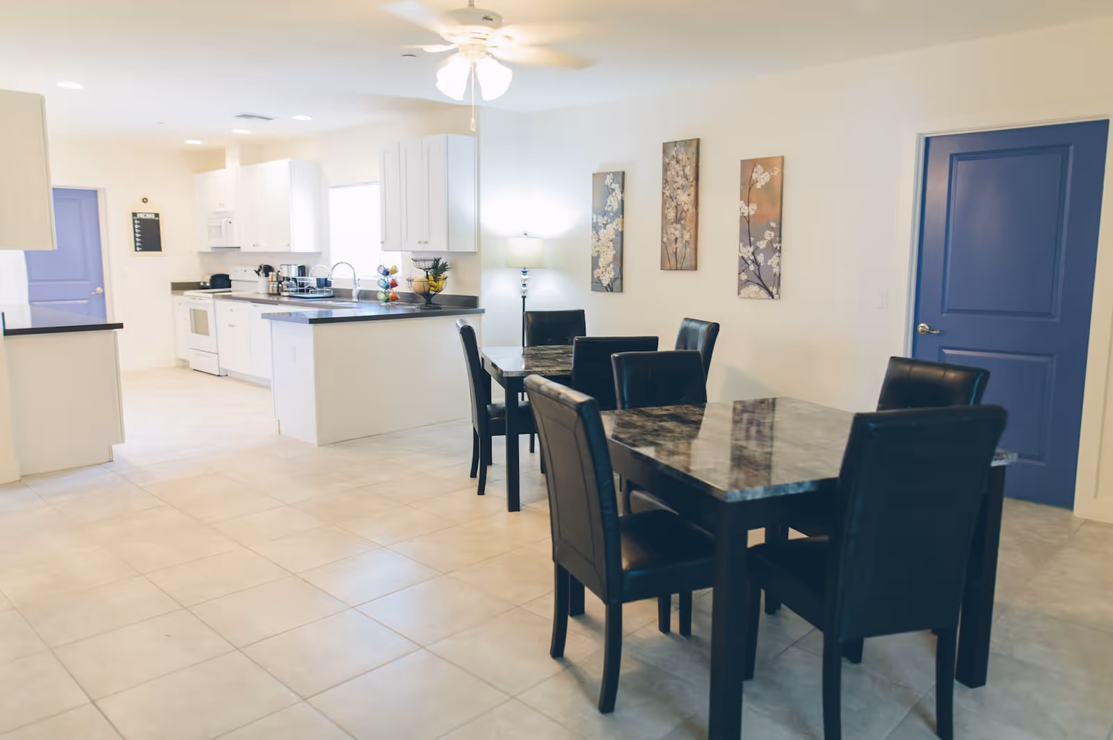 A bright and clean kitchen and dining area with white cabinets, black countertops, and tiled floors. The dining area features two dark marble tables with black leather chairs. Three floral paintings hang on the wall, and there is a ceiling fan with lights above.