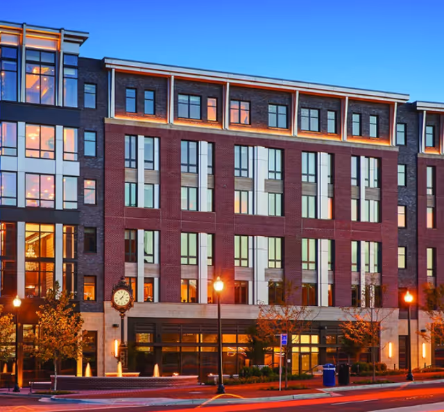 Exterior view of a modern multi-story building with large windows and brick facade during dusk. Street lamps and a decorative clock are visible in front of the building along with some small trees and a sidewalk.