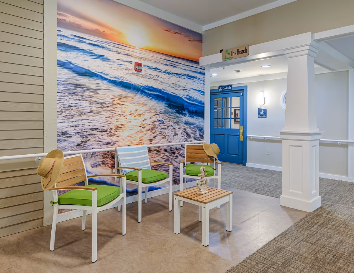 Indoor seating area with three white chairs featuring green cushions and a small wooden table. Two chairs have straw hats placed on them. The wall behind the chairs has a large mural of a beach sunset with waves. A blue door labeled 'Dockside' is visible in the background, along with a sign above the seating area that reads 'The Beach'.