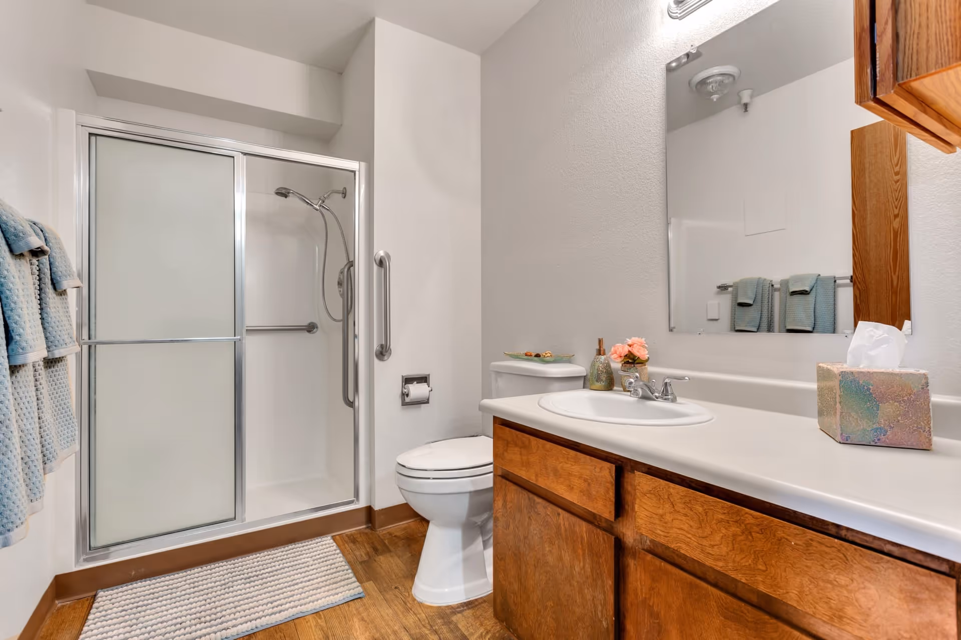 A clean bathroom with a walk-in shower featuring sliding frosted glass doors and a handheld showerhead. There is a white toilet next to the shower, a wooden vanity with a white countertop and sink, a large mirror above the sink, and a tissue box on the counter. Blue towels hang on a rack to the left, and a small rug is placed on the wooden floor in front of the shower.
