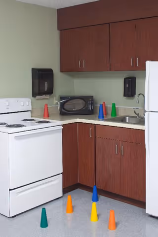 Small kitchen area with a white stove and refrigerator, wood cabinets, a microwave and sink, and colorful plastic cones on the counter and floor.