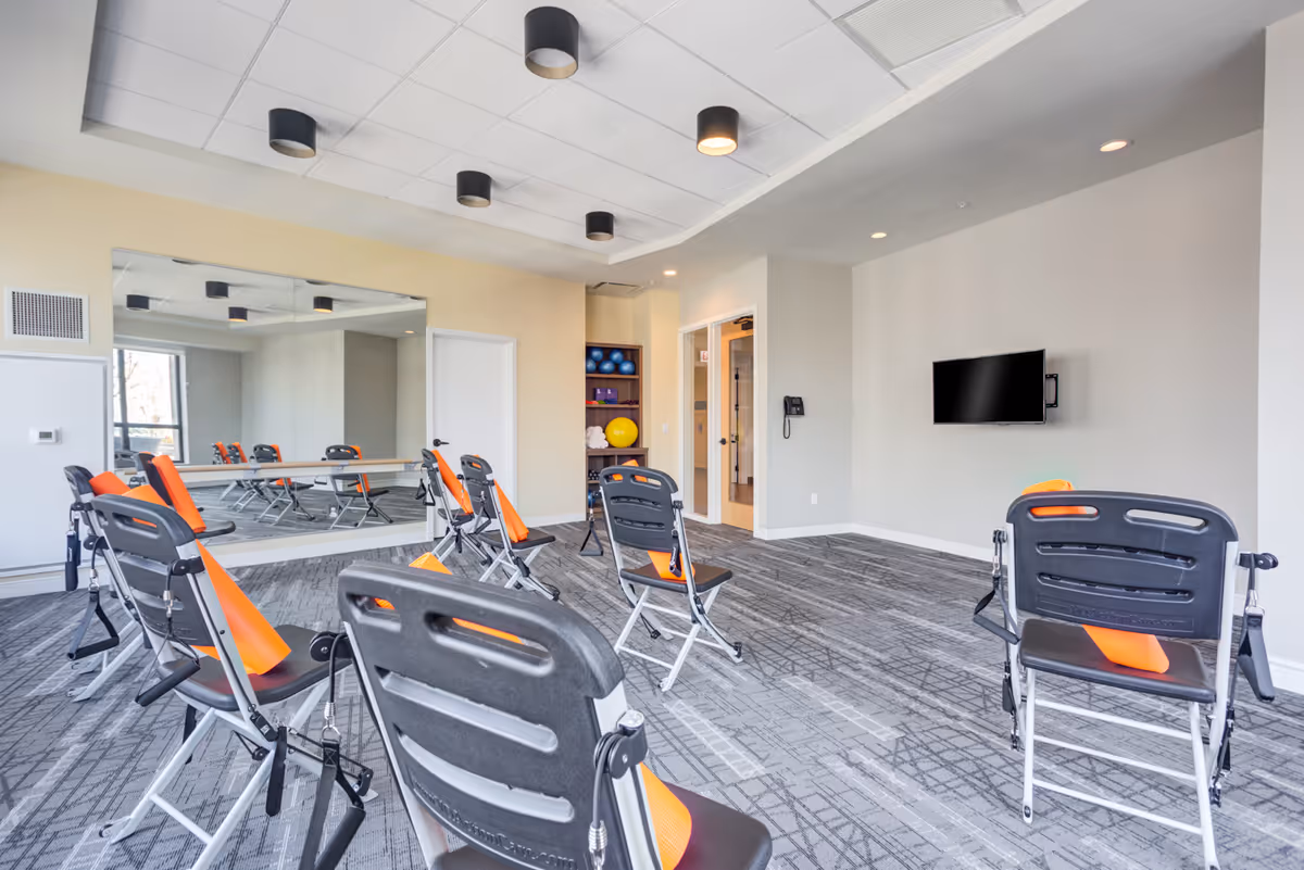 A bright exercise room with several black and orange chairs arranged in a semi-circle facing a wall-mounted TV. The room has a large mirror on one wall, a shelf with exercise balls and towels, and a gray patterned carpet. The walls are painted light beige and the ceiling has recessed lighting.