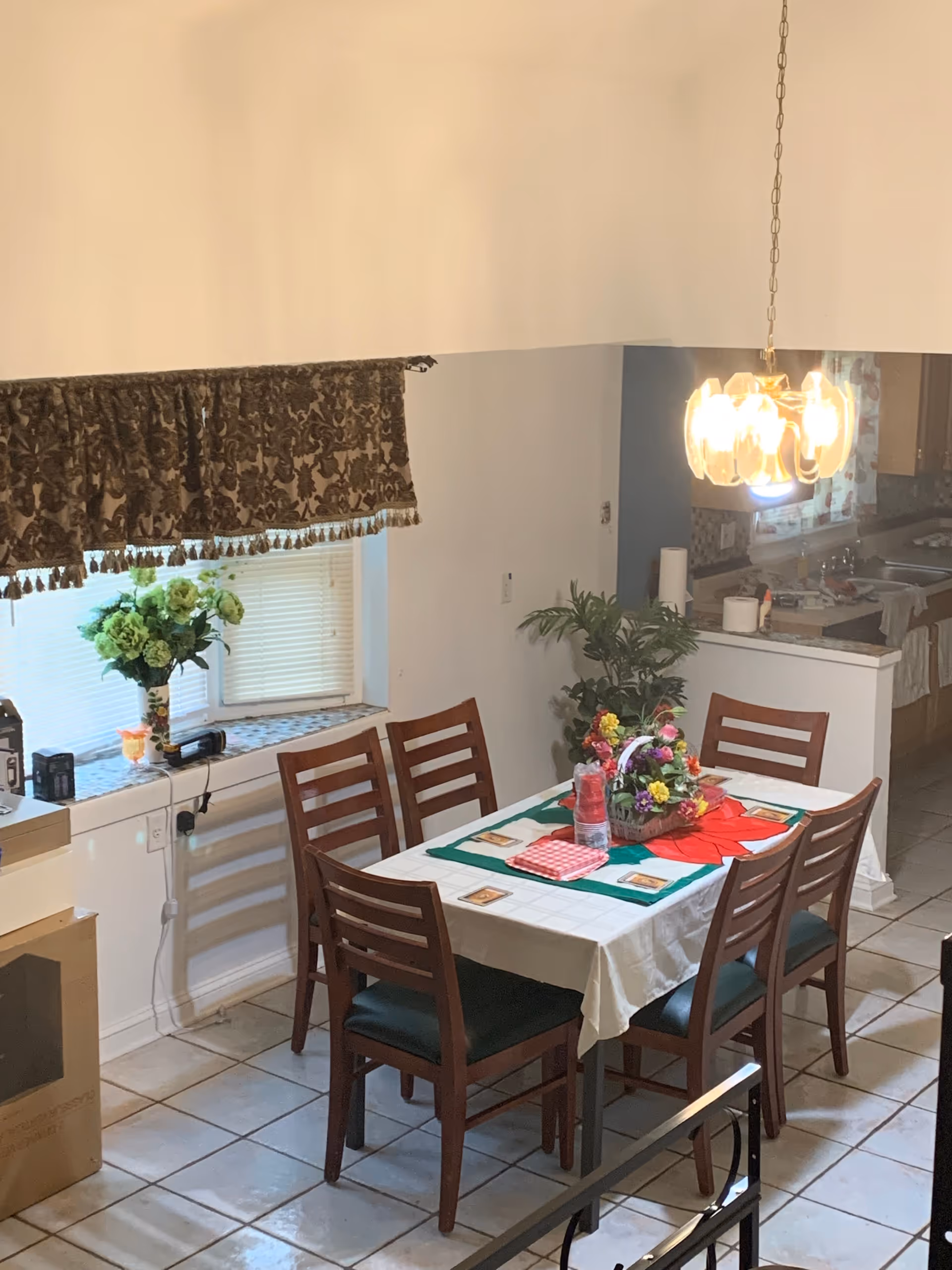 A dining area with a rectangular table covered with a white tablecloth and a green and red centerpiece. Six wooden chairs with dark cushions surround the table. A hanging light fixture illuminates the area. There is a window with a decorative valance and a vase of flowers on the windowsill. The dining area is adjacent to a kitchen with visible countertops and cabinets.