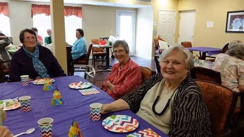 A group of elderly women sitting around a table covered with a purple tablecloth, decorated with colorful party hats and paper plates, in a communal dining area with yellow walls and windows with red valances.