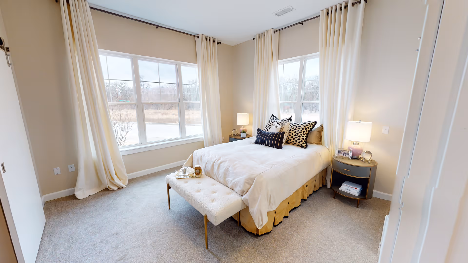 A bright and cozy bedroom with large windows covered by cream-colored curtains. The room features a neatly made bed with white bedding and multiple decorative pillows. There is a cushioned bench at the foot of the bed, and two round bedside tables with lamps and small decorative items. The carpet is light gray, and the walls are painted a soft beige.