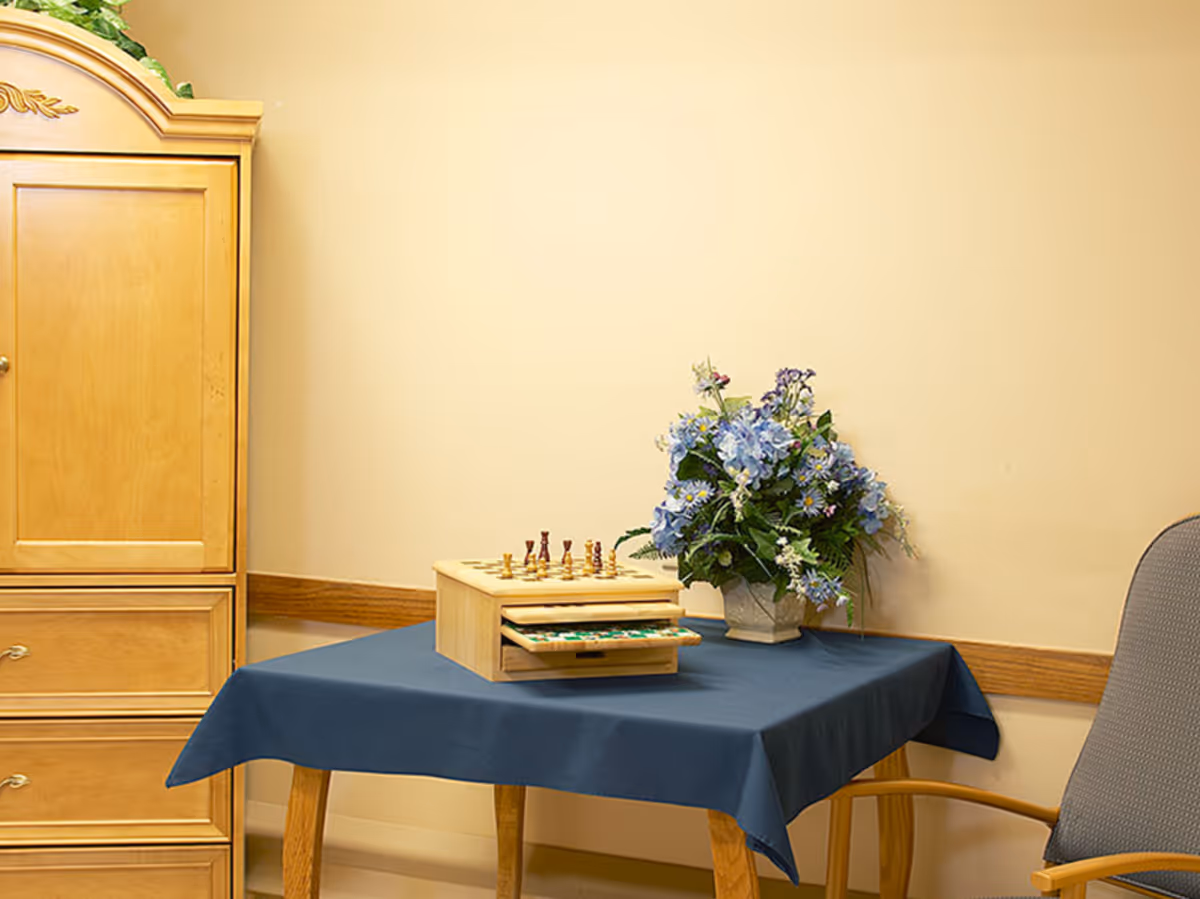 A small wooden table covered with a dark blue tablecloth, holding a wooden chess and checkers set and a vase with blue and white flowers. To the left is a wooden cabinet, and to the right is a gray cushioned chair with wooden armrests. The walls are painted a light beige color.