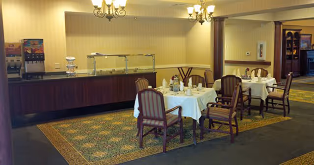 A dining area with two tables covered with white tablecloths, each set with plates, cups, and napkins. There are wooden chairs with striped upholstery around the tables. In the background, there is a buffet counter with a beverage dispenser and a food serving area. The room has warm lighting and carpeted floors with a patterned rug.