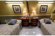 A shared bedroom in a senior living facility with two single beds separated by a nightstand. Each bed has a patterned bedspread and a pillow. The nightstand holds two lamps and a telephone. Above each bed hangs a framed floral artwork. The room has a warm, cozy ambiance with soft lighting.