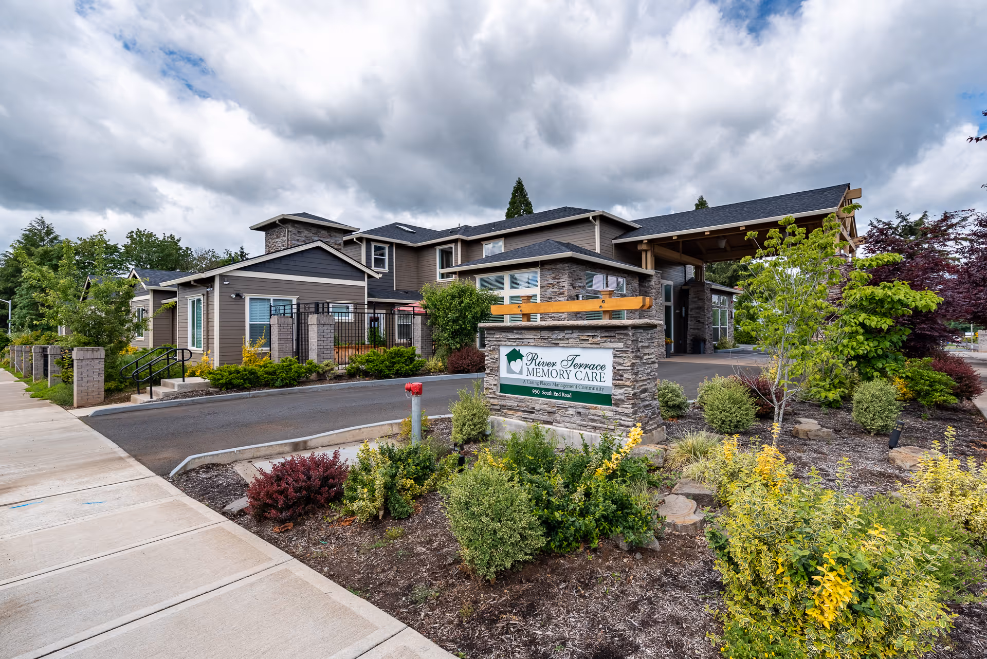 Front entrance of River Terrace Memory Care with a stone sign, landscaped gardens, and a covered driveway under a cloudy sky.