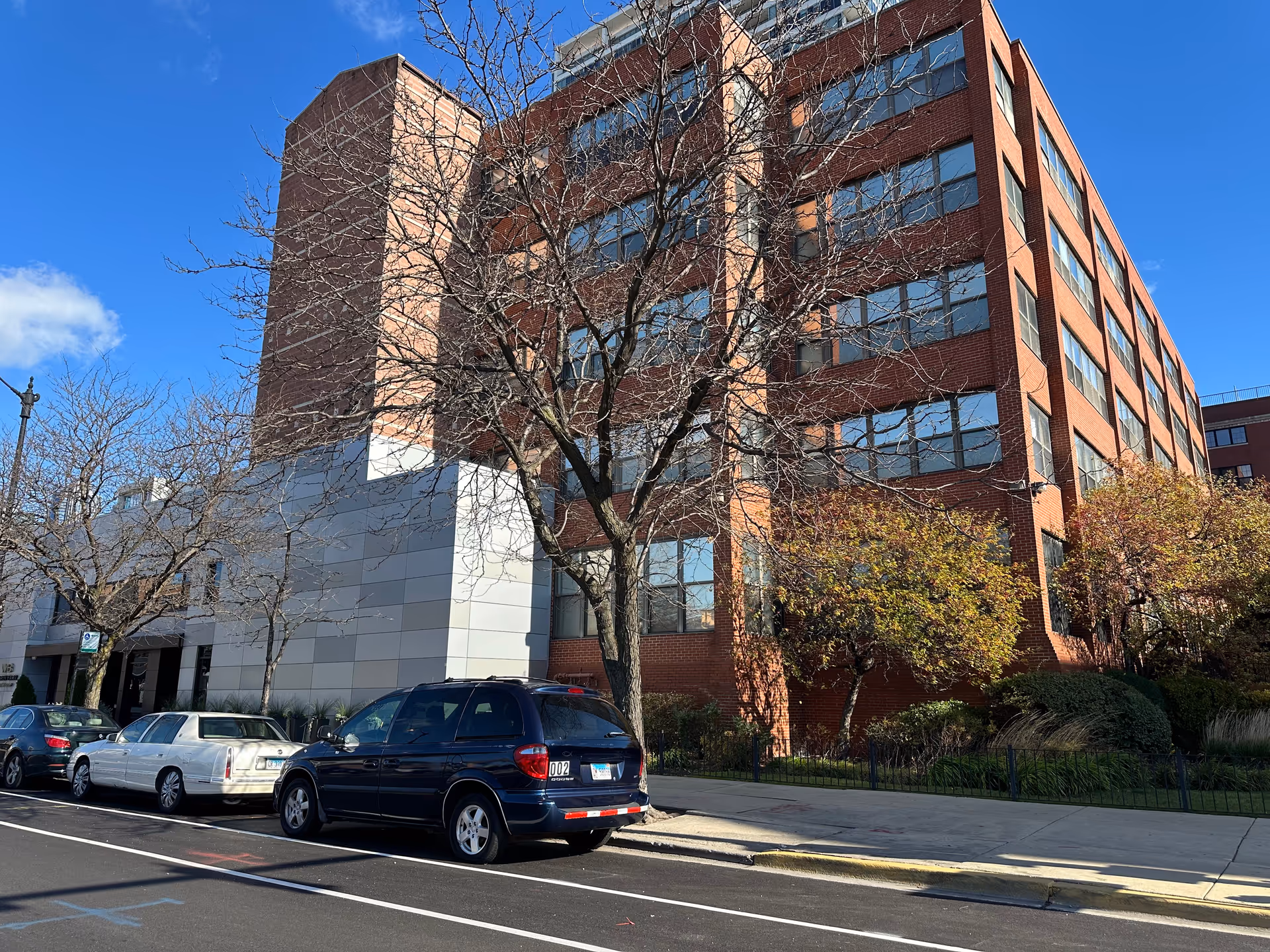 Street view of a multi-story red-brick building with parked cars, leafless trees, and a clear blue sky.