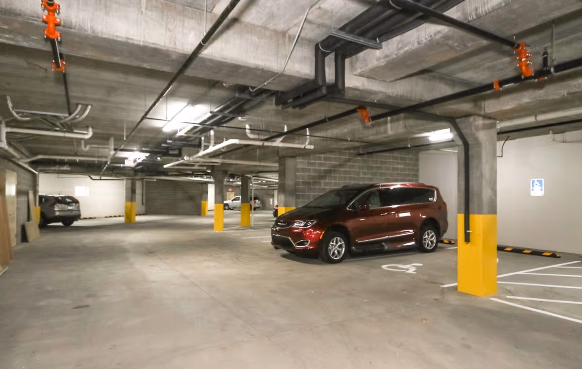 Indoor parking garage with concrete floors, ceiling, and pillars painted yellow at the base. Several cars are parked, including a red minivan in a handicapped parking space. Overhead pipes and lighting fixtures are visible on the ceiling.