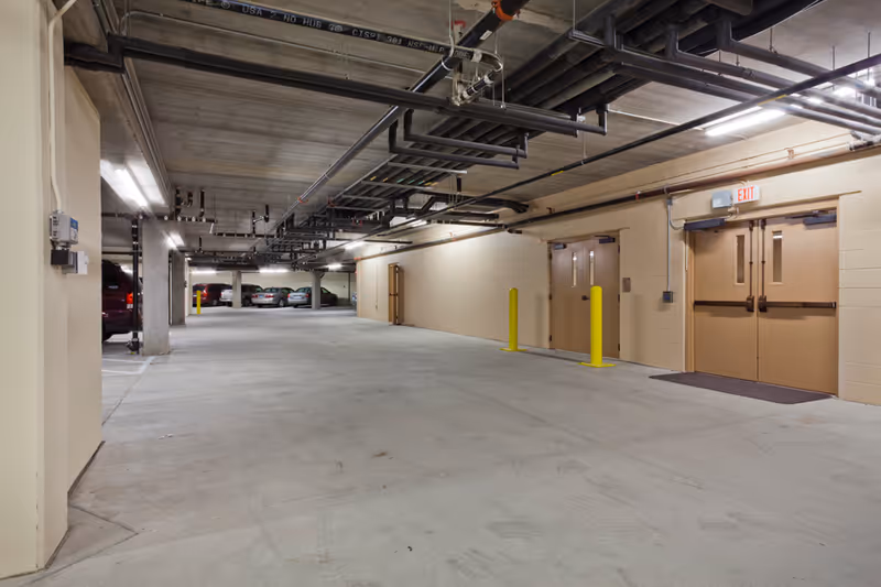 Underground parking garage with concrete floor and ceiling pipes, parked cars in the distance, and beige double doors marked with an exit sign.