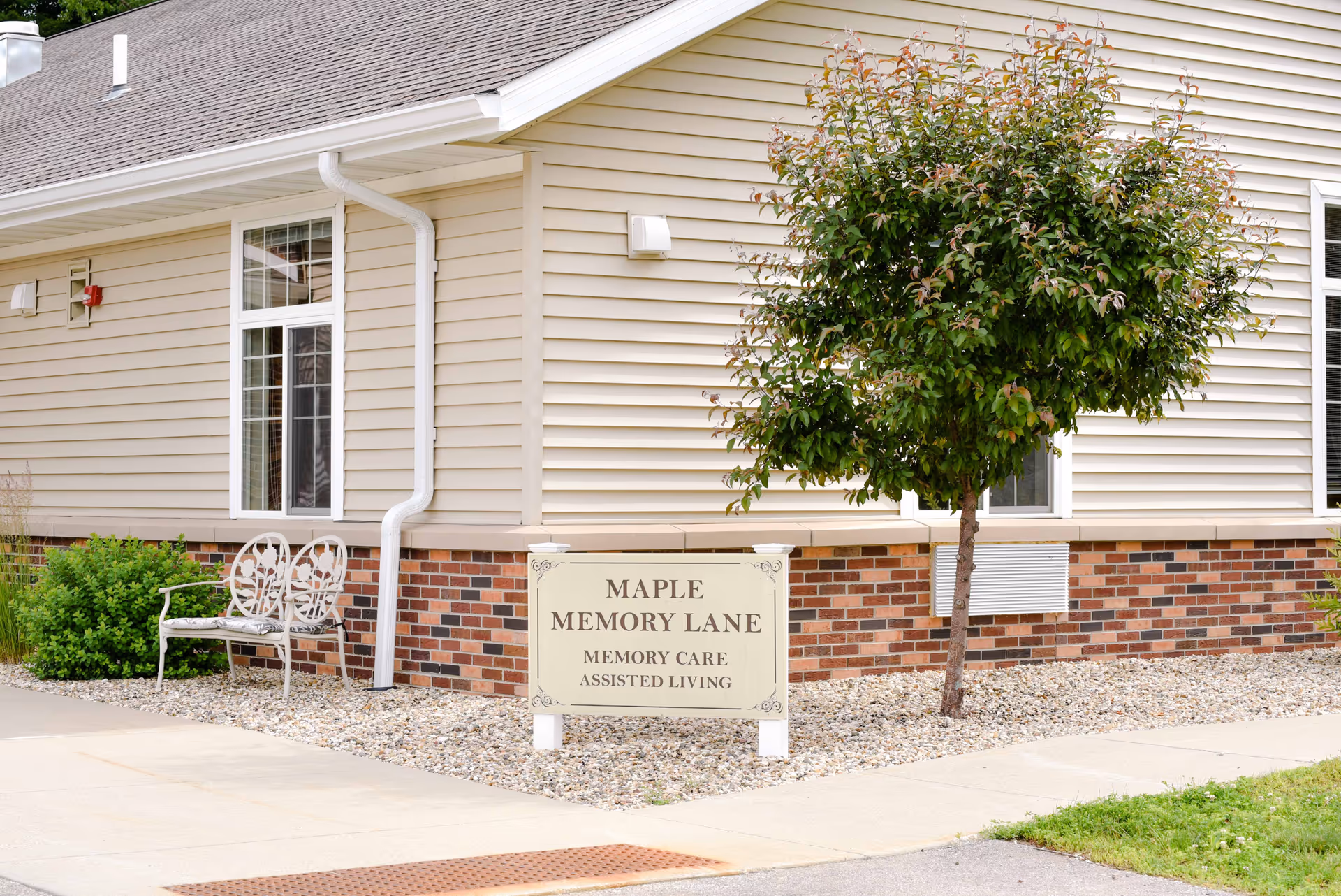 Exterior view of a beige building with brick accents, a small tree, a white bench, and a sign that reads 'Maple Memory Lane Memory Care Assisted Living'.