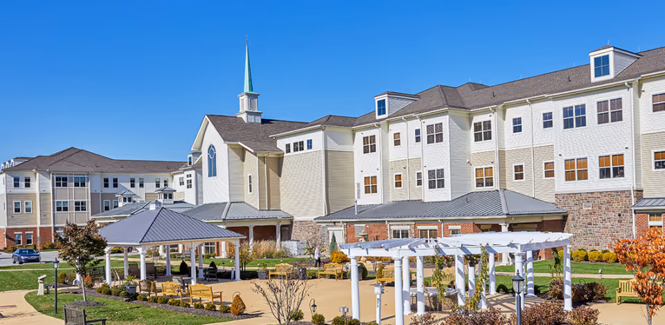 Exterior front view of a multi-story senior living building with a landscaped courtyard, pergola, gazebo, and benches under a clear blue sky.