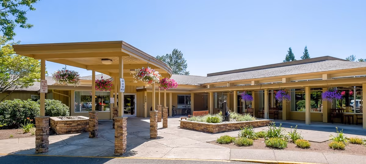 Covered entrance and porte-cochere of a single-story senior living building with hanging flower baskets and a small landscaped courtyard.