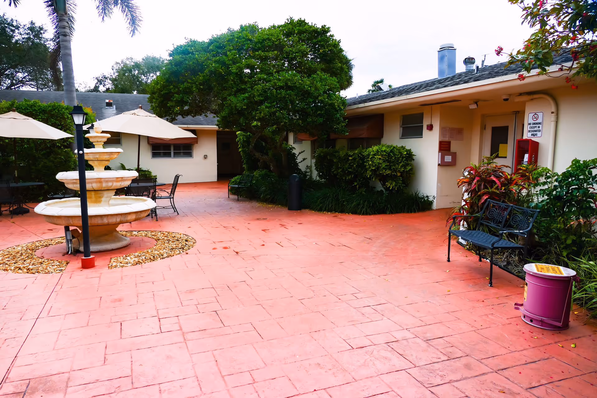 Outdoor courtyard area with a three-tiered fountain surrounded by a circular bed of small rocks, several tables with umbrellas, chairs, benches, and lush greenery including trees and bushes. The courtyard is paved with reddish tiles and is adjacent to a single-story building with a door and windows.