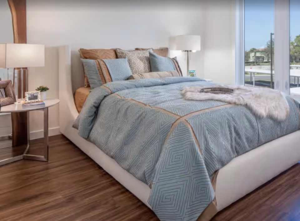 A neatly made bed with a light blue and beige patterned comforter and multiple pillows in a bright bedroom with wooden flooring. There is a round side table with a lamp, a small plant, and a photo frame next to the bed. Large windows on the right side allow natural light to fill the room, showing an outdoor view.