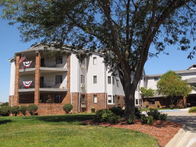 Exterior view of a multi-story senior living building with brick lower levels, balconies decorated with bunting, and a large tree on the lawn.