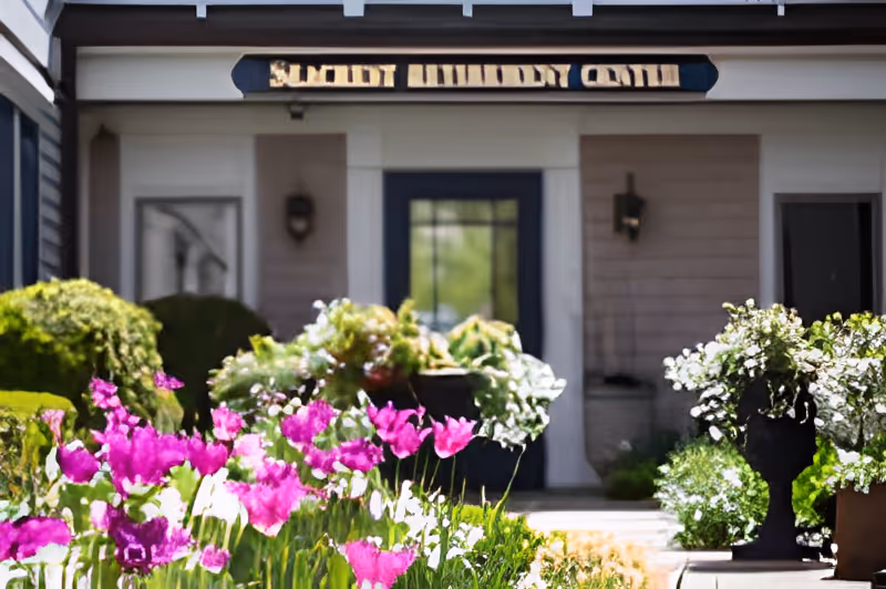 Entrance to a building with a sign above the door reading 'Seacrest Assisted Living & Memory Care,' surrounded by vibrant pink and white flowers and green shrubs.