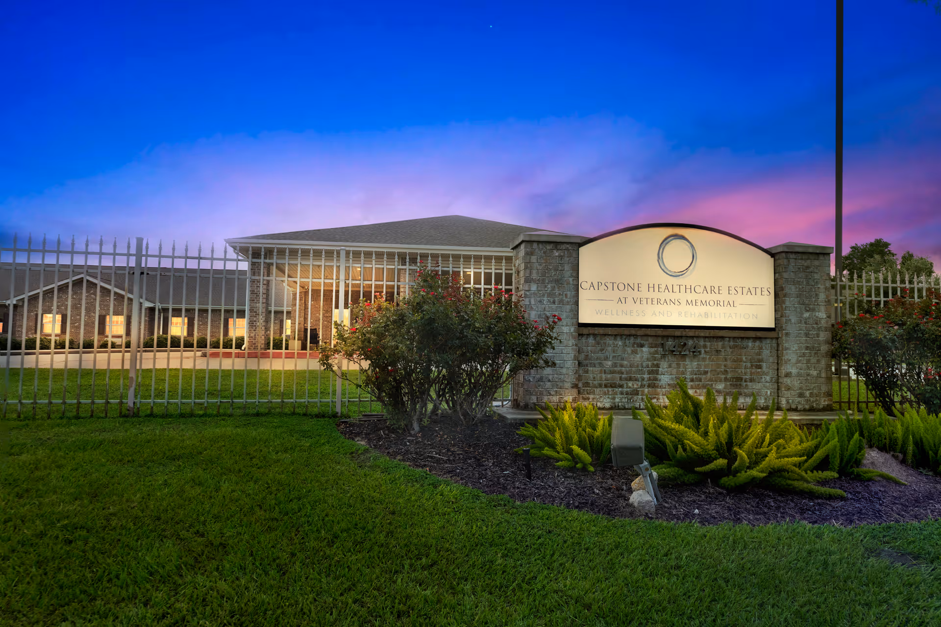 Exterior view of Capstone Healthcare Estates at Veterans Memorial building during twilight, showing a brick sign with the facility name, surrounded by a metal fence, green grass, and landscaping with bushes and plants under a vibrant blue and purple sky.