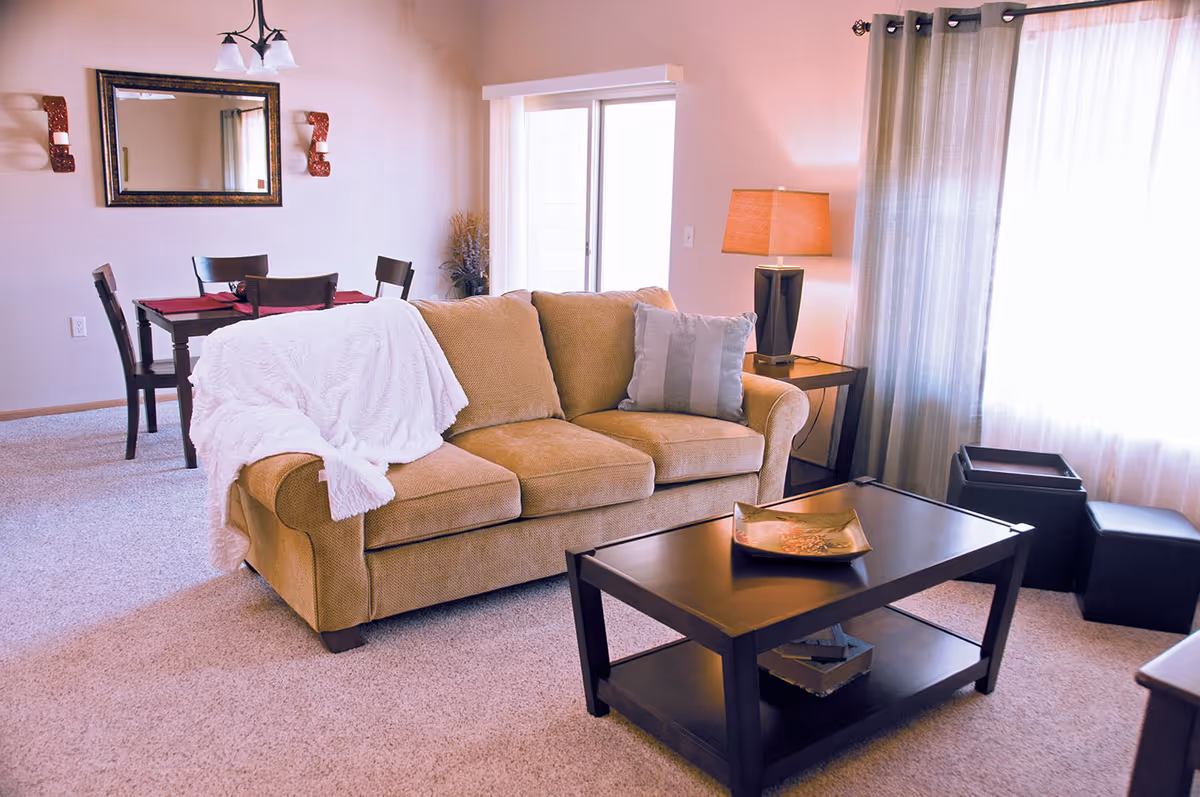 A cozy living room with a beige sofa draped with a white blanket and a gray pillow. In front of the sofa is a dark wooden coffee table with a decorative tray. To the right, there is a side table with a lamp and large windows with curtains letting in natural light. In the background, a dining area with a wooden table and four chairs is visible, along with a wall mirror and two wall sconces.