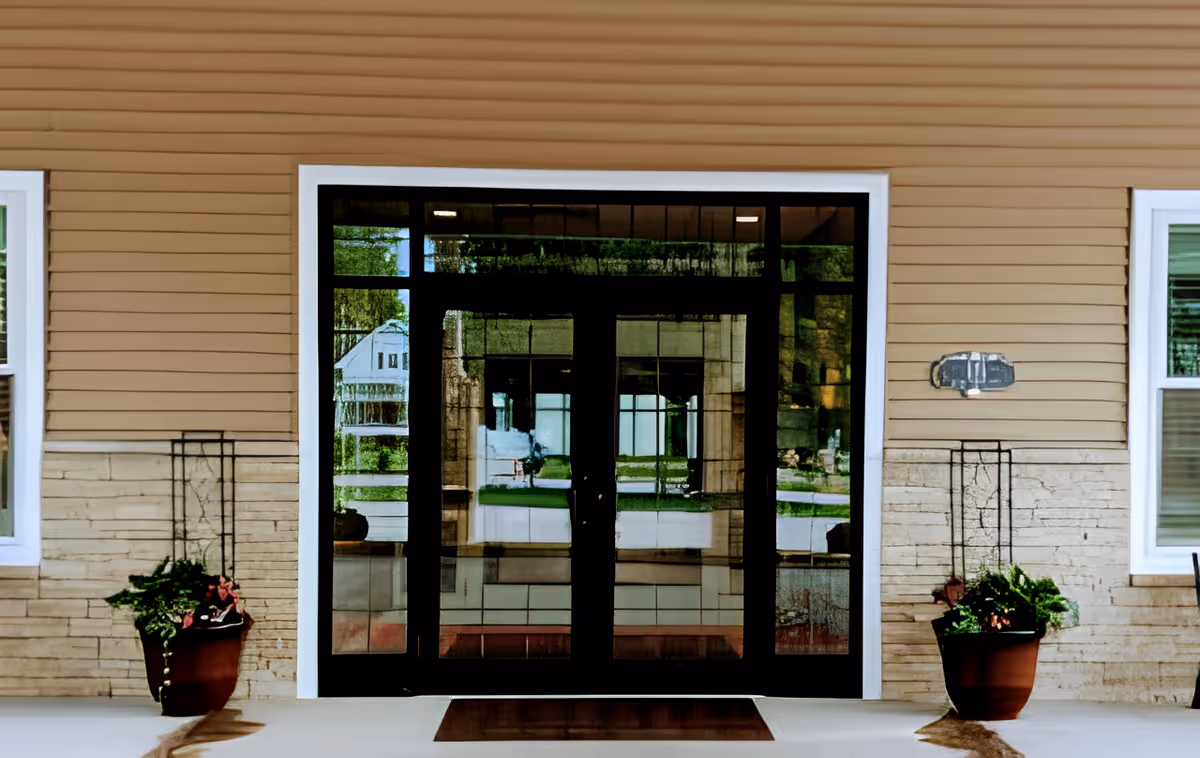 Glass double doors entrance to a building with beige siding and stone lower walls, flanked by two large potted plants with greenery and flowers, and two windows on either side.