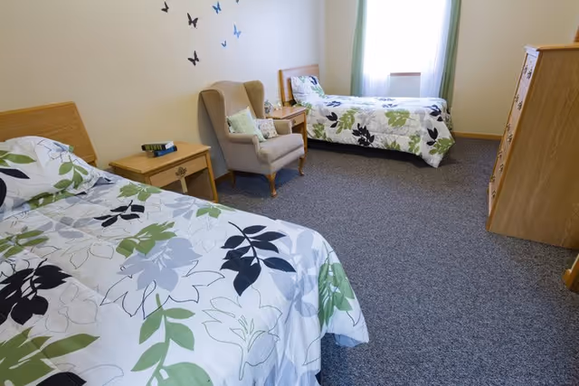 A simple bedroom with two twin beds covered in floral patterned bedspreads in shades of green, black, and gray. Between the beds is a wooden nightstand with a few books on it. There is a beige upholstered armchair with a cushion next to the nightstand. The room has a window with white curtains and green drapes, and a wooden dresser is visible on the right side. The walls are light-colored with butterfly decorations above the armchair.