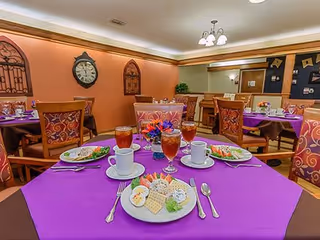 Dining room with tables covered in purple tablecloths, set with plates of food, cups, glasses of iced tea, and silverware. The room has warm lighting, decorative wall clocks, and framed wall decor.
