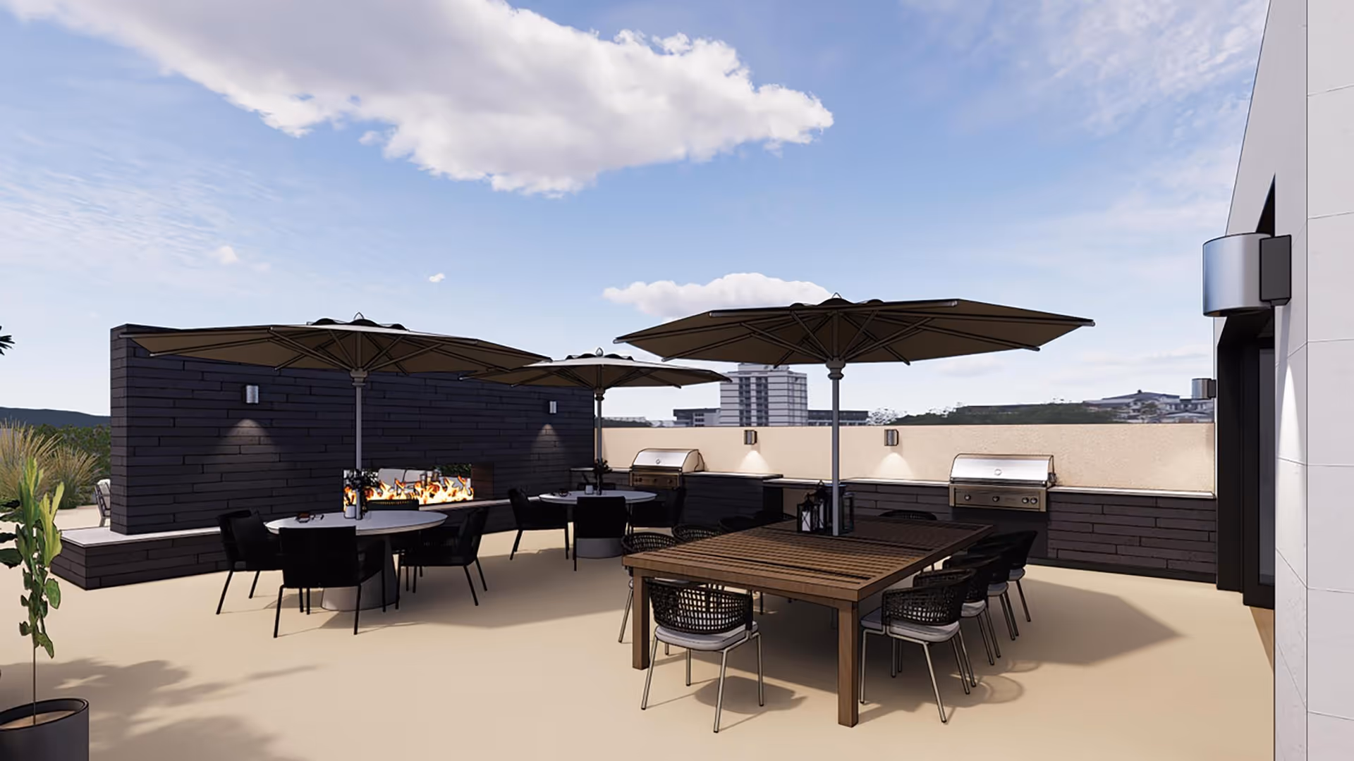 Outdoor patio area with multiple tables and chairs under large umbrellas. There are built-in grills and a modern fire pit against a dark brick wall. The sky is partly cloudy with a few buildings visible in the background.
