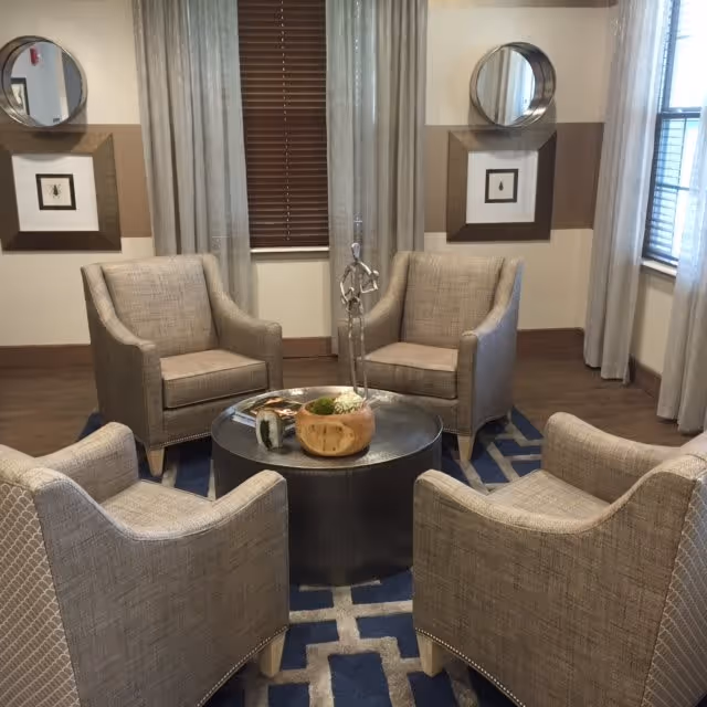 A cozy seating area with four beige upholstered armchairs arranged around a round black coffee table. The table has decorative items including a wooden bowl and a small sculpture. The room features a blue and beige patterned rug, two round mirrors on the wall, framed artwork, and windows with blinds and light curtains.