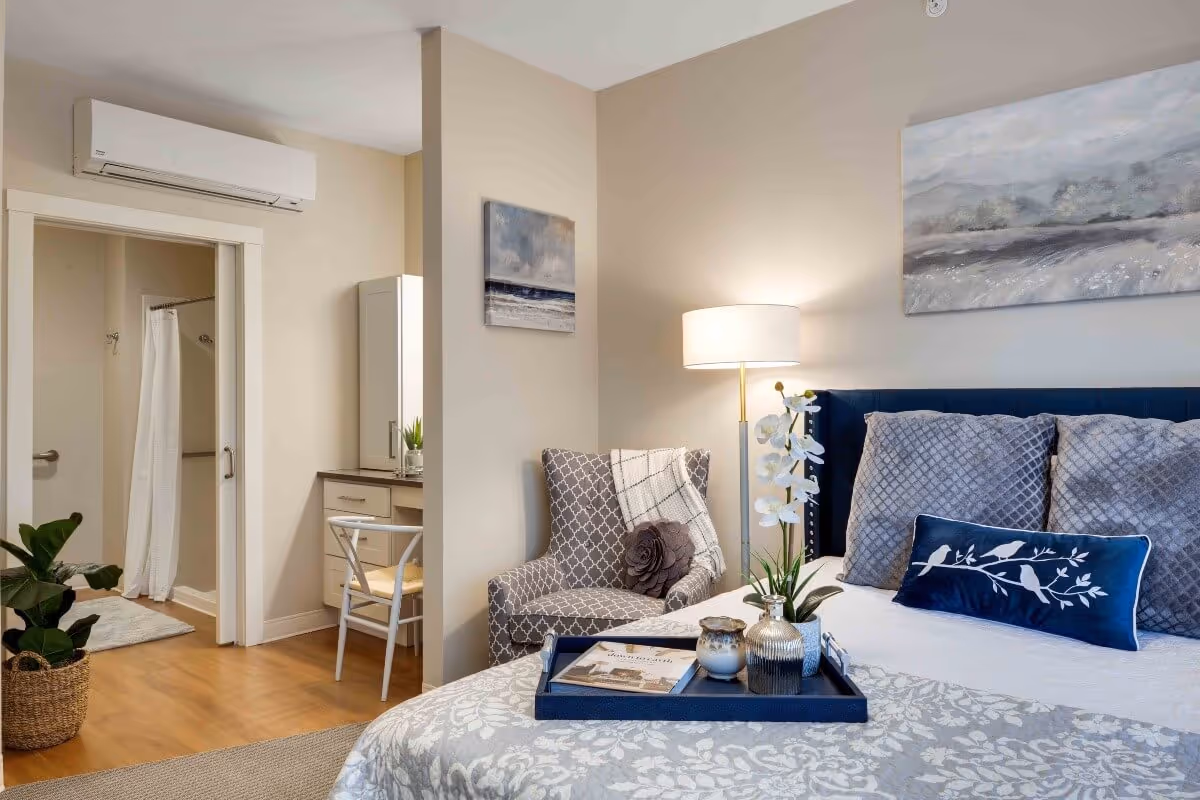 A cozy bedroom in a senior living facility featuring a bed with gray and blue pillows, a patterned armchair with a throw blanket and decorative pillow, a floor lamp, and a small vanity desk with a chair. The room has light-colored walls and wood flooring. A bathroom with a shower and white curtain is visible through an open door. There is a potted plant on the floor near the bathroom entrance.