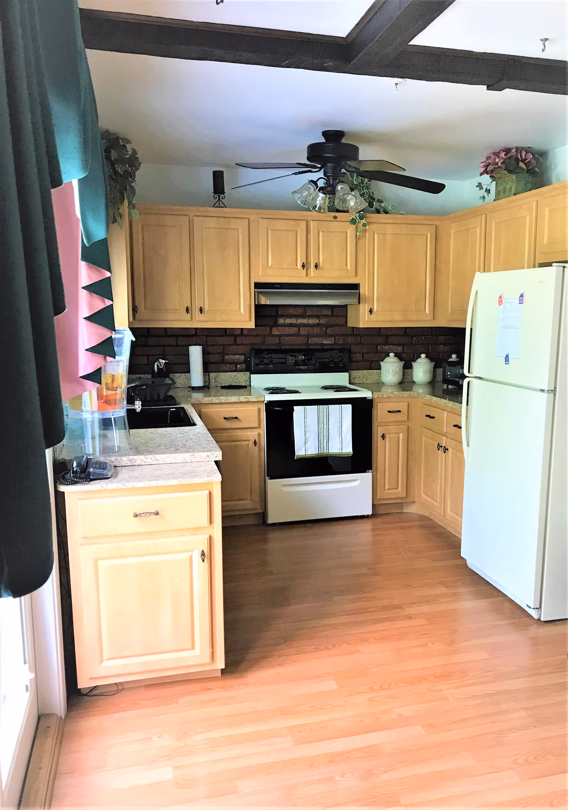 A kitchen with light wood cabinets, a white refrigerator, a white stove with a black range hood, and a countertop with a sink. There is a ceiling fan with lights, decorative plants on top of the cabinets, and a water dispenser on the counter near a window with green and pink curtains.