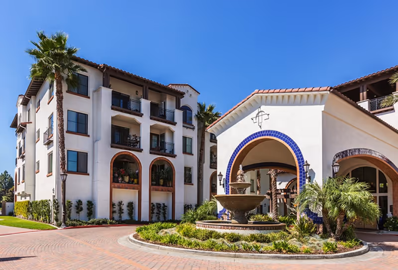 A senior living facility with white stucco buildings, arches, palm trees, and a central fountain.