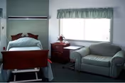 A nursing home bedroom with a hospital-style bed, wooden nightstand, window with green valance, armchair, and small table with flowers.