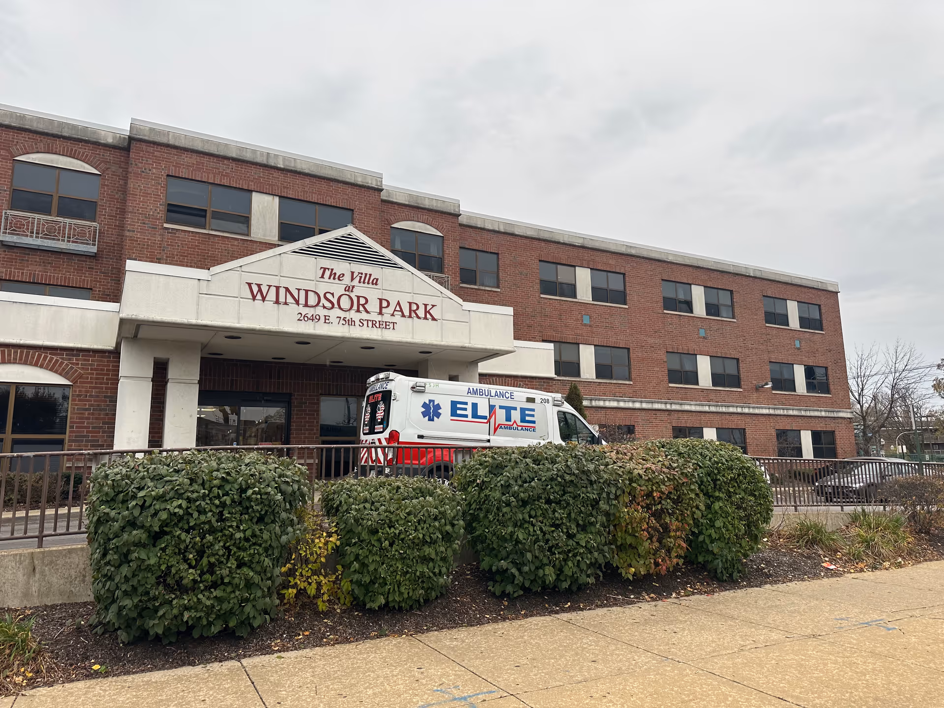 Exterior view of a three-story brick building with a sign reading 'The Villa at Windsor Park 2649 E. 75th Street' above the entrance. An ambulance with 'Elite Ambulance' written on the side is parked in front of the building. There are trimmed bushes and a sidewalk in the foreground under a cloudy sky.