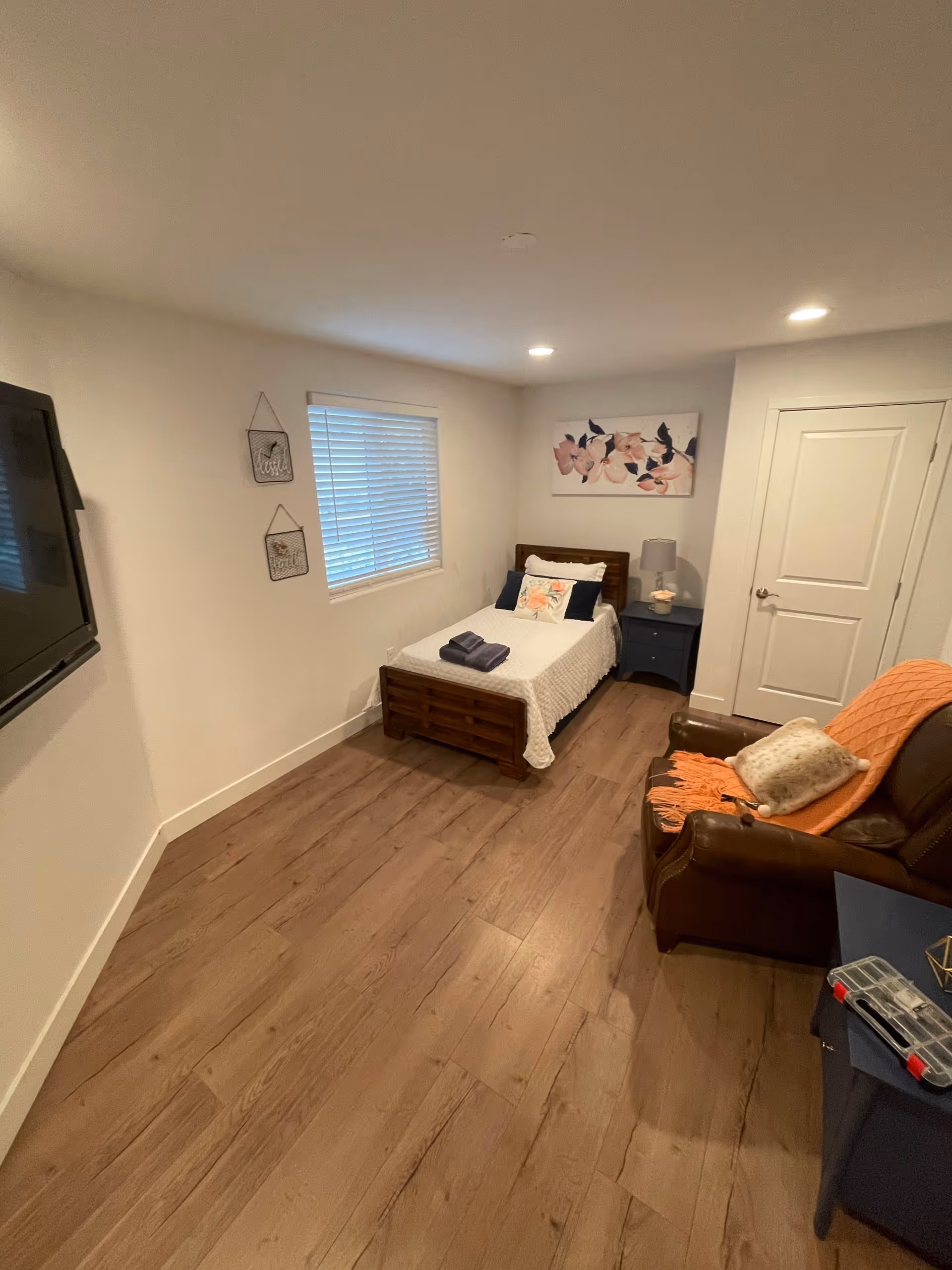 A cozy bedroom with a single wooden bed covered with a white quilt and decorative pillows. A folded towel is placed on the bed. Next to the bed is a blue nightstand with a lamp and a small decorative item. A brown leather armchair with an orange throw blanket and a pillow is positioned near the nightstand. The room has wooden flooring, a window with blinds, and a flat-screen TV mounted on the wall. There are two small framed wall hangings and a floral painting above the bed. A closed white door is visible in the background.
