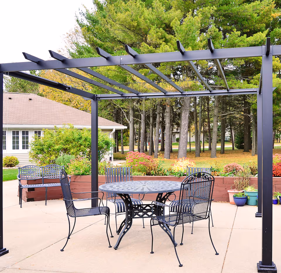Outdoor patio with a metal pergola, round metal table and chairs, benches and potted plants in front of trees and a nearby building.
