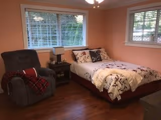 A cozy bedroom with peach-colored walls, a bed with floral bedding, a gray recliner chair with a red and black plaid blanket draped over it, a small dark wooden nightstand with a lamp, and two windows with white blinds letting in natural light.