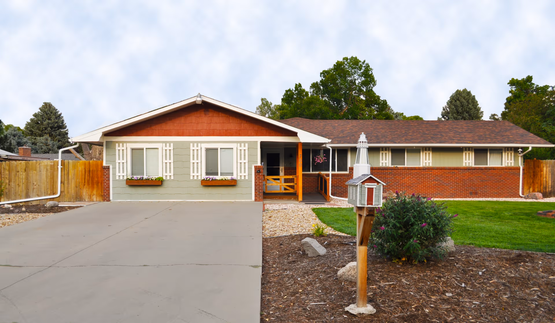 Single-story ranch-style house front with a driveway, small lawn, flower boxes, and a decorative mailbox shaped like a lighthouse.