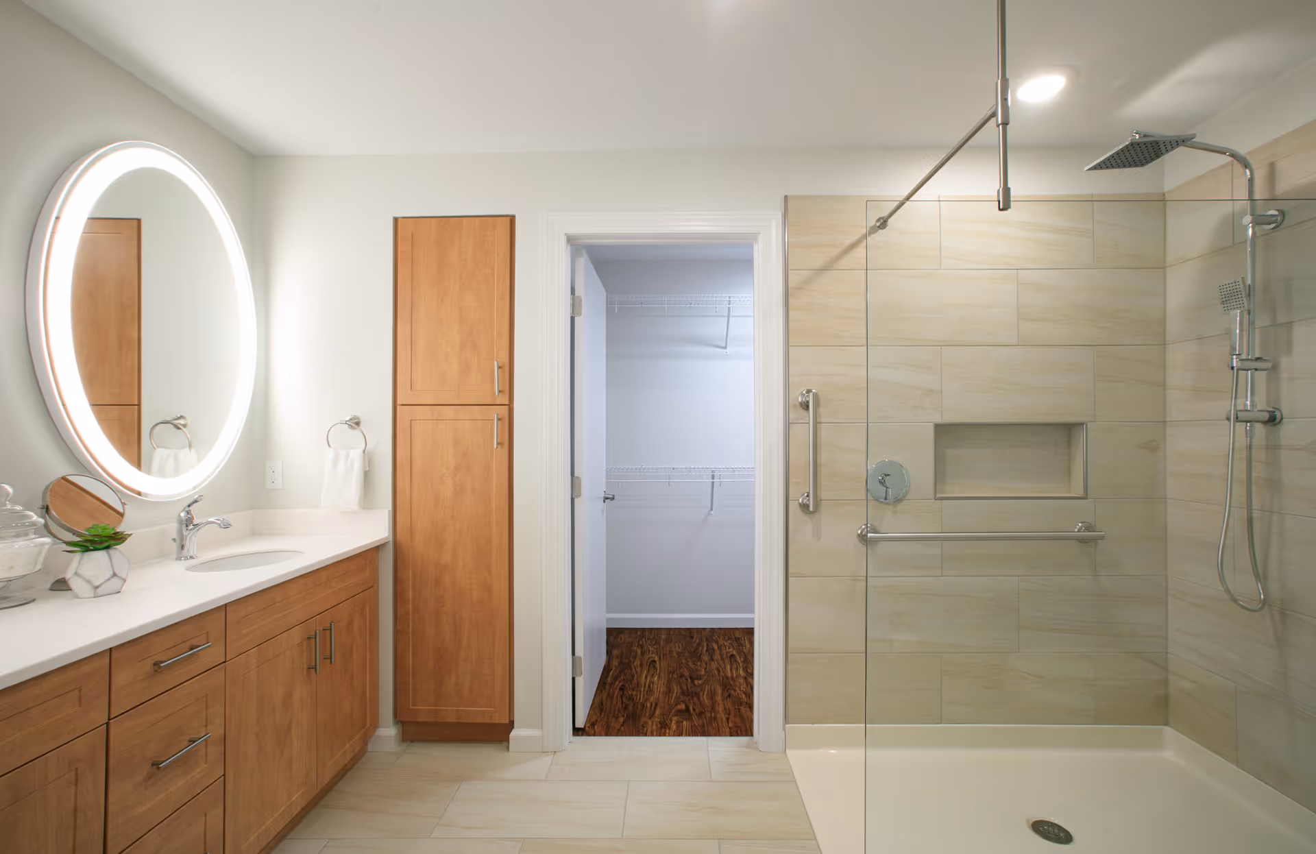 Modern bathroom with light wood cabinetry, a round illuminated mirror above a white countertop with a sink, a glass-enclosed shower with beige tiles, and a doorway leading to a walk-in closet with wooden flooring and white wire shelving.